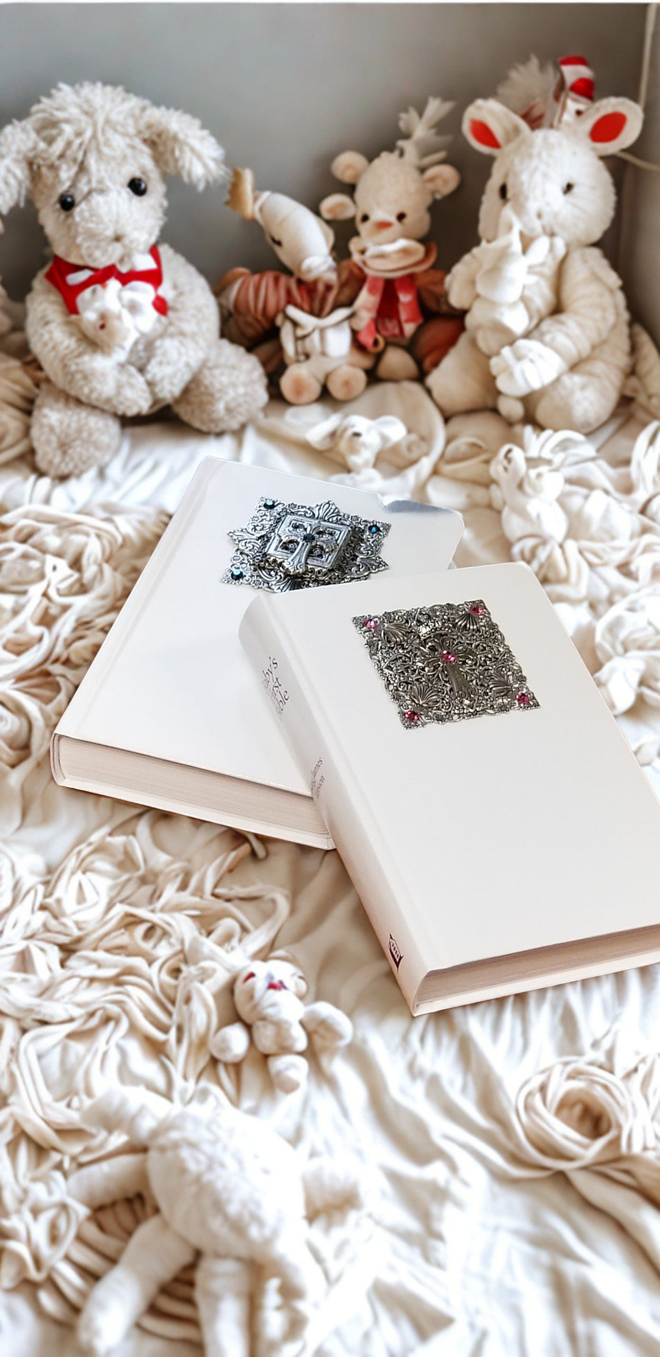 White baby Bibles with ornate cross covers, nestled among plush toys on a cozy blanket