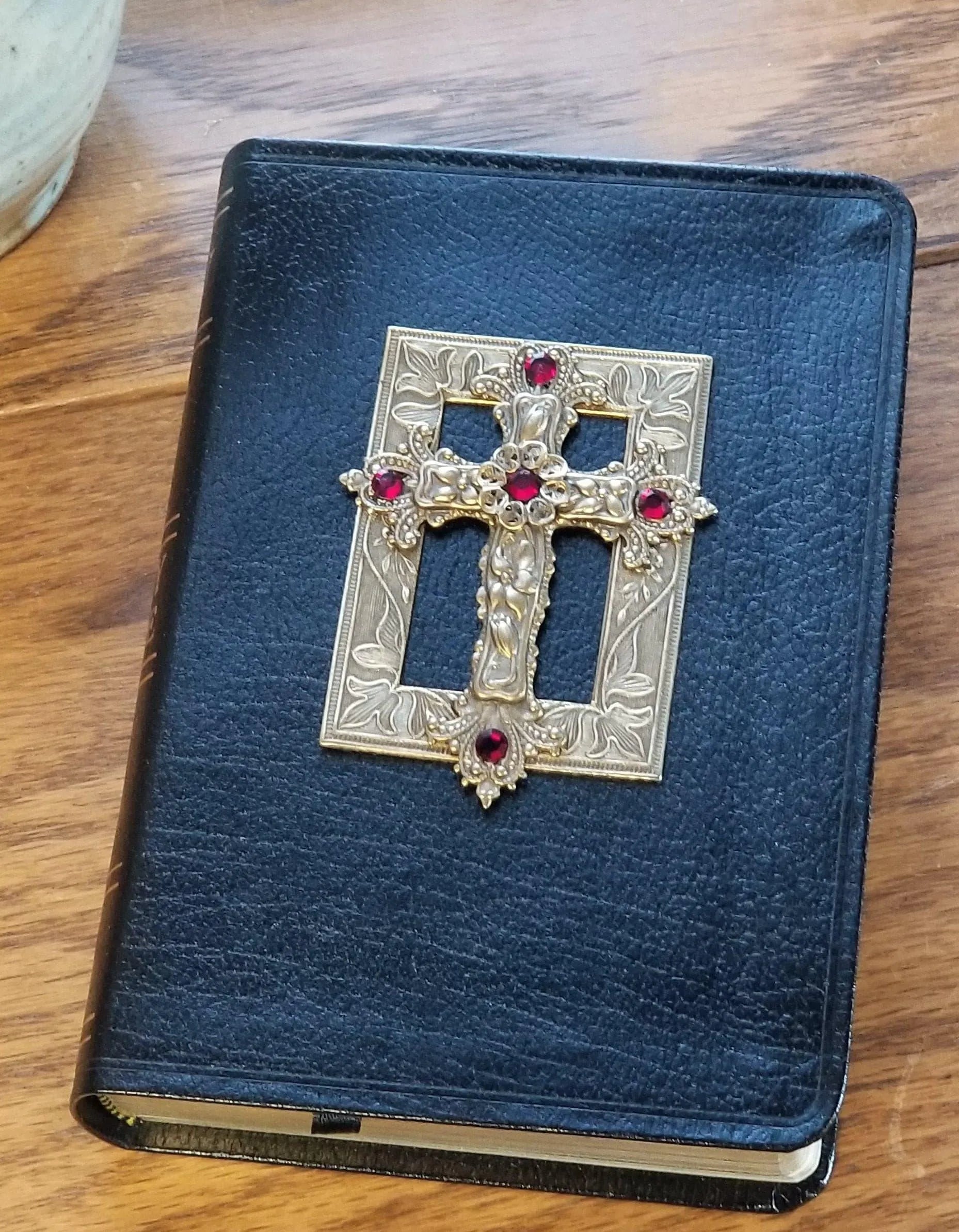 Black leather Bible with ornate gold cross and red jewel accents on wooden table