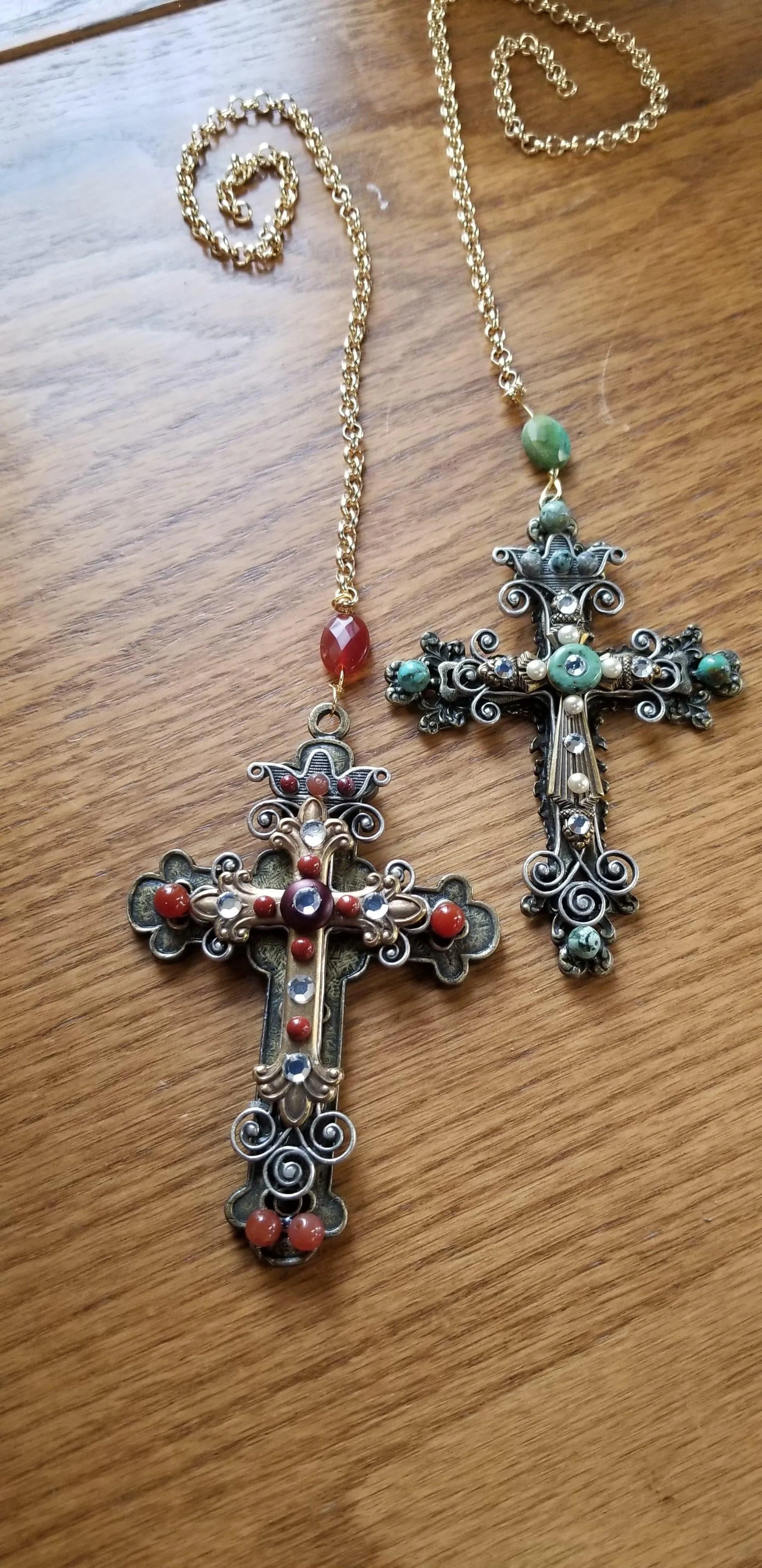 Jeweled cross ornaments with red, green, gold, and silver accents on gold chains, on wood surface.