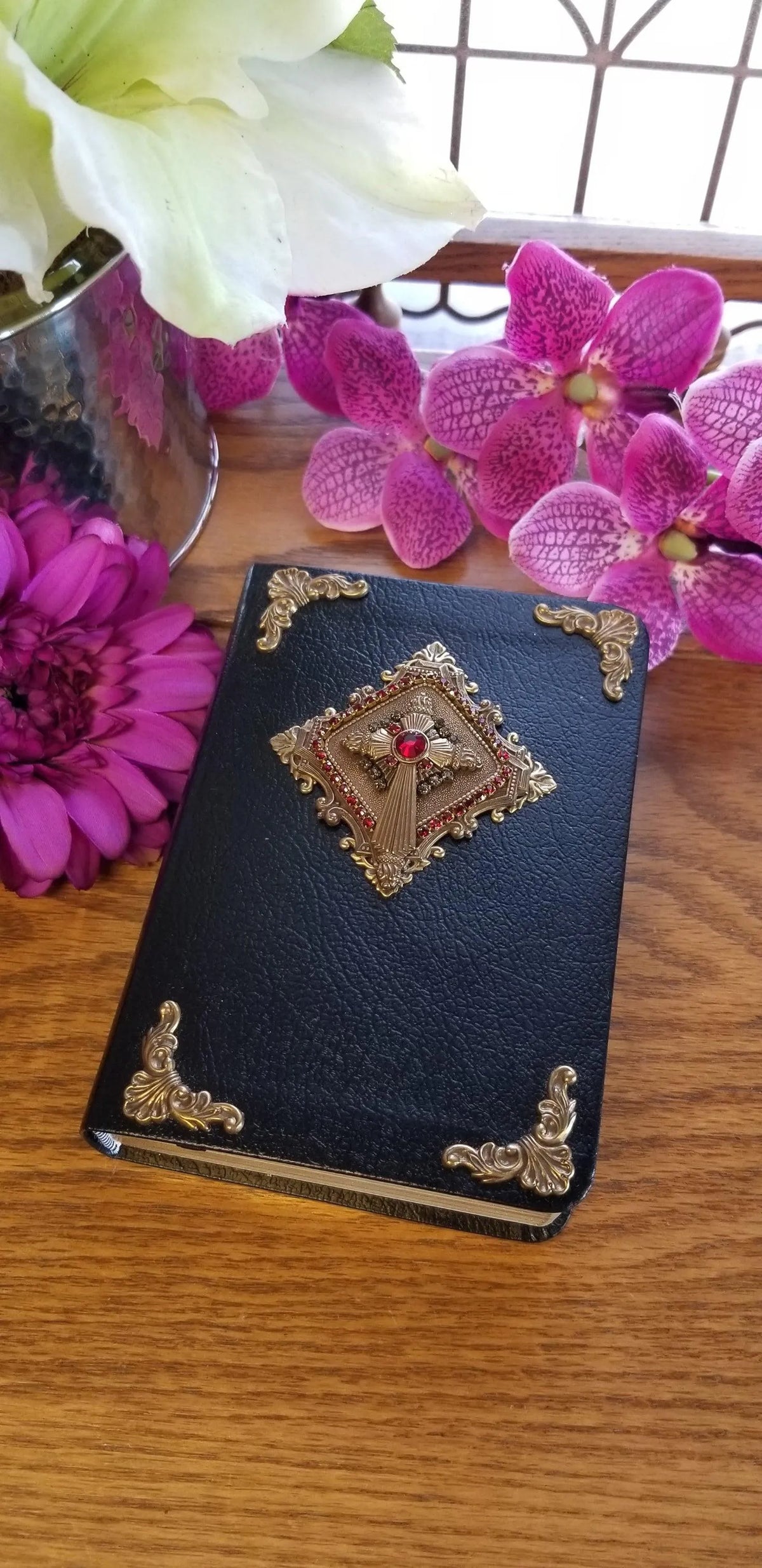 Jeweled heirloom Bible with black cover, gold accents, and red gem, surrounded by flowers.