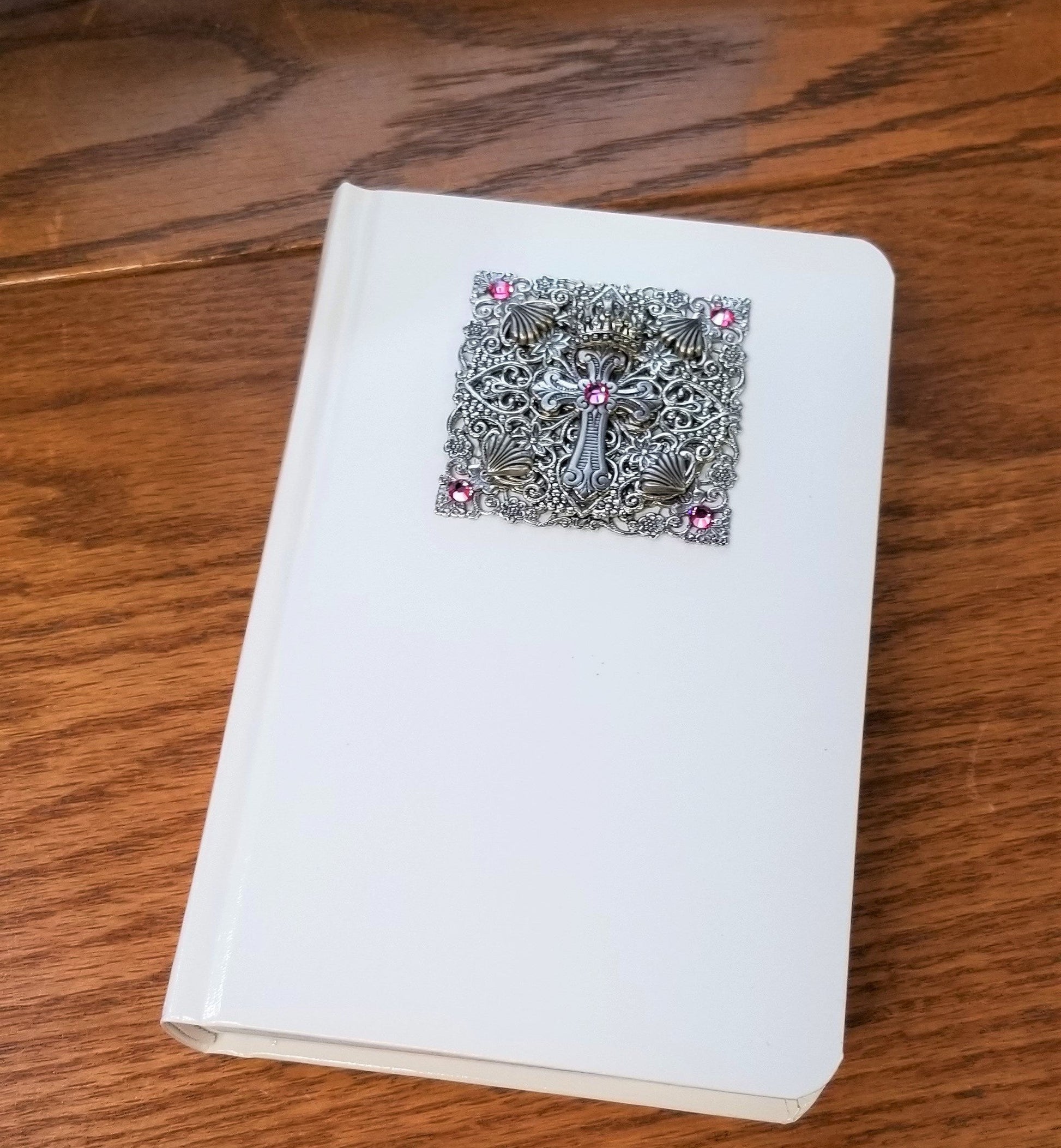 White baby Bible with ornate silver cross and pink jeweled accents on cover, on wood surface
