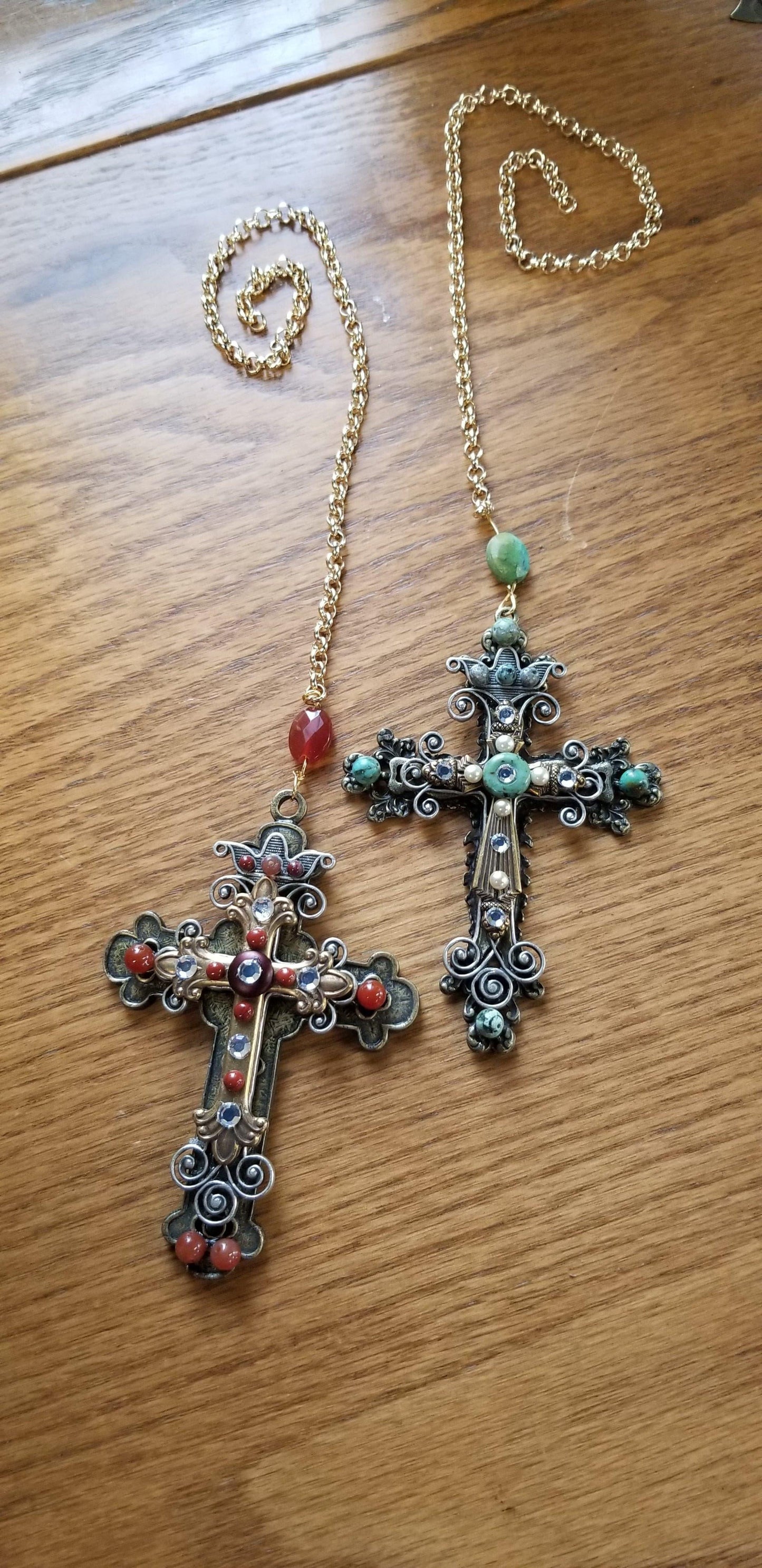 Pair of ornate cross ornaments with gold chains, jeweled with red and green stones on wood surface.